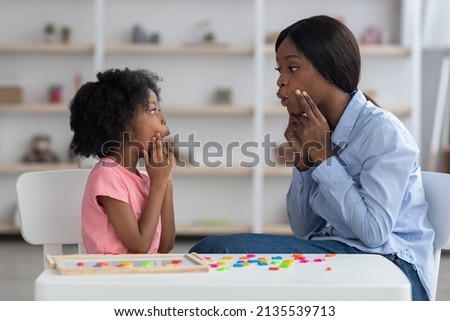Young african american woman speech-language pathologist having lesson with little girl with cute curly hair, black teacher and pupil working on pronunciation, touching faces and grimacing Royalty-Free Stock Photo #2135539713
