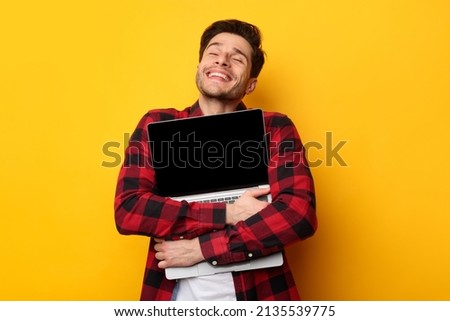 Technology Lover Concept. Happy man hugging laptop with black blank empty screen, holding it tight near chest. Casual male geek with closed eyes standing isolated on yellow orange studio wall Royalty-Free Stock Photo #2135539775