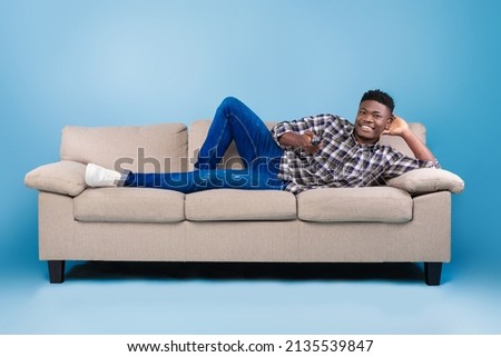 Weekend leisure activities. Millennial black man with remote control switching channels on TV while lying on couch over blue background. African American guy watching interesting program on television Royalty-Free Stock Photo #2135539847