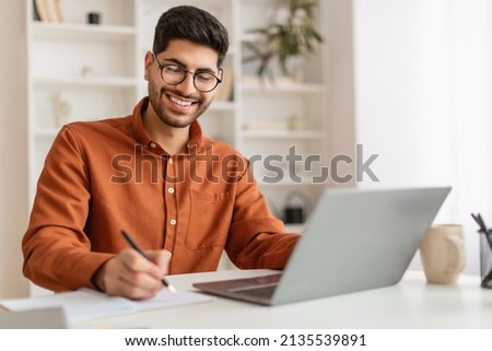 Portrait of young smiling Arabic man in glasses using laptop sitting at desk, writing in notebook. Happy guy working remotely, watching webinar training studying online, taking notes signing documents