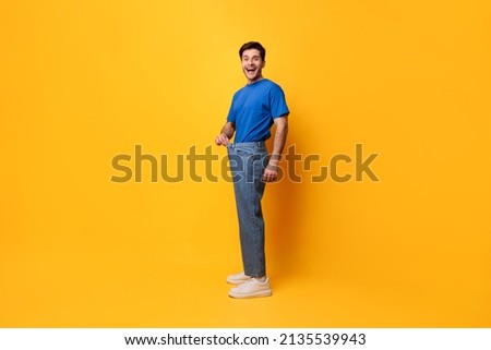 Successful Weight Loss. Happy Excited Slim Young Guy Pulling Old Large Loose Jeans Achieved Slimming Weightloss Goal Standing Posing Isolated On Yellow Studio Wall, Full Body Length, Free Copy Space Royalty-Free Stock Photo #2135539943