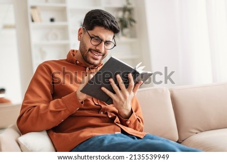 Portrait of young Middle Eastern man wearing eyeglasses holding paper book or diary, sitting on couch at home in living room, reading literature or checking his working schedule plan, free copy space Royalty-Free Stock Photo #2135539949