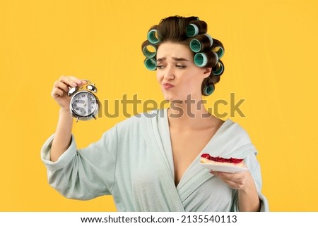 Diet. Hungry Glamorous Female Holding Cake And Clock Eating Sweets After 6 P.M. In The Evening Struggling To Lose Weight Standing Over Yellow Studio Background. Weight Loss, Nutrition Concept