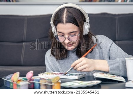 A stylish young woman artist in glasses and headphones draws with paints at home.