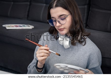 Stylish young woman in glasses, teenager, draws a picture on canvas while sitting near the sofa in the interior of the house.