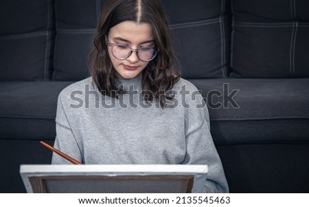 Stylish young woman in glasses, teenager, draws a picture on canvas while sitting near the sofa in the interior of the house, copy space.