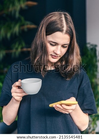 Teen girl with a cup of tea and a smartphone in her hands on a blurred dark background.