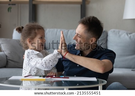 Adorable small laughing preschool child girl giving high five to joyful caring young daddy, having fun drawing picture in paper album, finishing entertaining domestic activity, daycare concept.
