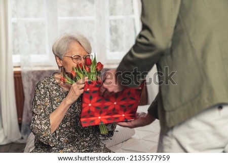 back view man giving his elderly grey-haired mother a bouquet of red tulips and a red paper bag present for Mother's Day Grandparent's Day. High quality photo