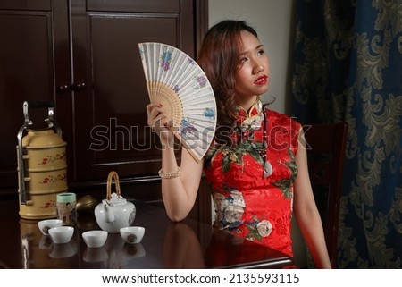 Attractive young woman wearing retro Chinese cheongsam dress sitting next to vintage table with tea set tiffin carrier chair looking holding checking posing paper hand fan