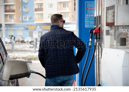 A man with a blue jacket and dark glasses at a gas station. He fills up the car. Petrol. Lifestile.