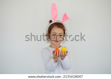 A little cute girl in rabbit ears holds colorful eggs in her hands. She is wearing a white T-shirt. Easter. Preparation for the holiday.