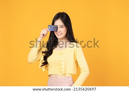 Cheerful young asian woman in yellow T-shirt holding bank card isolated on yellow background