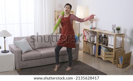 cheerful young wife wearing an apron is singing into the feather boom and shaking her body during housecleaning at home living room.