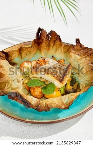 Baked halibut fillet with vegetables in parchment. Fish dish - roasted halibut with potato, broccoli and cauliflower. Food menu in minimal concept. Baked white fish on white concrete background