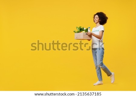Full body young smiling woman of African American ethnicity wear white volunteer t-shirt walk going hold food box isolated on plain yellow background. Voluntary free work assistance help grace concept