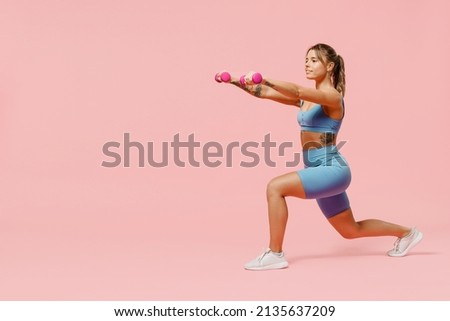 Full body young sporty athletic fitness trainer instructor woman wear blue tracksuit spend time in home gym hold dumbbells do stretch squats isolated on plain pink background. Workout sport concept.