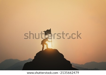 Business Success, Leadership and Success and Goal Concepts. Silhouette of businessman with flags on mountain peaks above sky and sunset background. Royalty-Free Stock Photo #2135662479