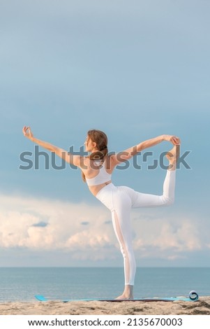 Young fit woman in white sportswear practices yoga. Natarajasana pose on the beach on bright sunny day