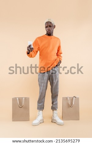full length of african american man in orange sweatshirt taking selfie on smartphone near shopping bags on beige