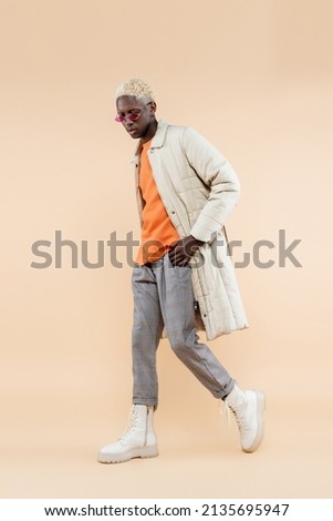 full length of young african american man in stylish sunglasses and coat posing with hand in pocket on beige Royalty-Free Stock Photo #2135695947