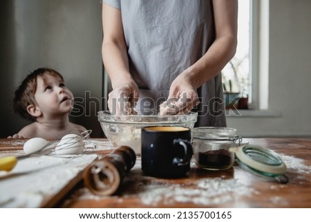 A woman in a gray apron prepares dough in a transparent bowl on a wooden table. A child stands next to her and watches the process. There are foods on the table. Only hands in focus
