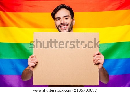 Portrait of happy smiling homosexual man holding empty blank board, with a gay pride flag behind him at studio. Lgbtq flag, rainbow flag, celebrating parade.