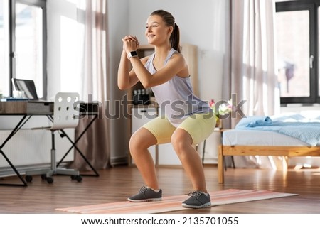 sport, fitness and healthy lifestyle concept - smiling teenage girl with smart watch exercising on yoga mat and doing squats at home Royalty-Free Stock Photo #2135701055