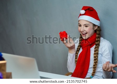 Expressive Positive Smiling Caucasian Girl Wearing Santa Hat Using Laptop for Online Video Virtual Chat While Holding And Demonstrating Red Giftbox on Laptop Device at Home. Horizontal Image