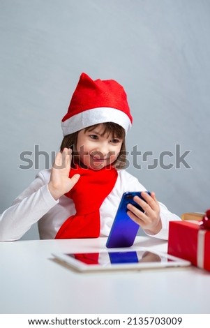 Smiling Caucasian Girl In Santa Hat Using Cellphone and Digital Pad For Chatting and Online Video Classroom With Waving Hand and Saying Hello From Home. Vertical Image