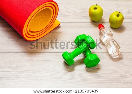 Yoga mat, apples, a bottle of water and dumbbells in a room or a gym on the gray floor.