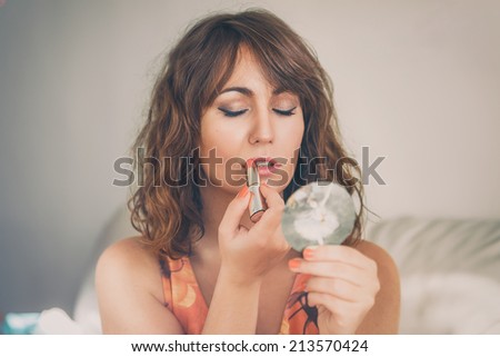 Attractive young woman with tousled brunette hair applying her lipstick using a handheld mirror in a fashion and beauty concept