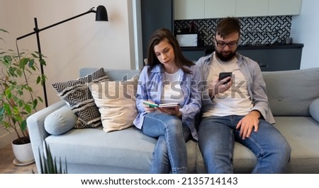 Young couple in love in casual outfits in white t-shirts mocap in jeans sit on the couch boyfriend uses his phone girlfriend reads her book. Concept: living together using gadgets smartphone phone 
