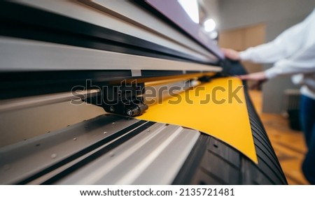 Female hands holding yellow printable material on alarge format printing plotter. Graphic design and  advertising concept. Royalty-Free Stock Photo #2135721481