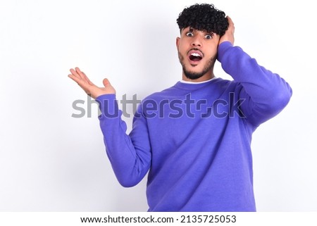 Shocked amazed surprised young arab man with curly hair wearing purple sweatshirt over white background hold hand offering proposition