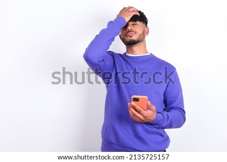 Upset depressed young arab man with curly hair wearing purple sweatshirt over white background makes face palm as forgot about something important holds mobile phone expresses sorrow and regret blames Royalty-Free Stock Photo #2135725157