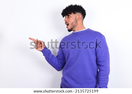 Stunned young arab man with curly hair wearing purple sweatshirt over white background with greatly surprised expression points away on copy space, indicates something