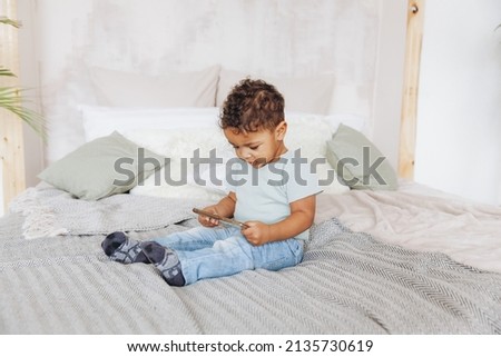 Cute little african american boy with smartphone at home on bed, happy kid watching cartoons on phone.
