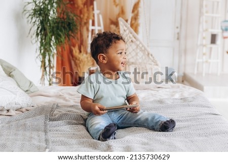 Cute little african american boy with smartphone at home on bed, happy kid watching cartoons on phone.