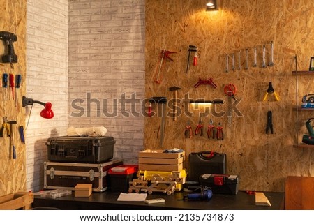 Artisan woodwork studio with shelving holding instruments, tools for carpentry and repair works. Empty workshop with Screwdriver, pliers and hammer hanging on board, workbench and toolbox. Royalty-Free Stock Photo #2135734873