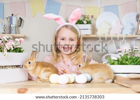 
Easter little girl with rabbit ears holds 2 live rabbits in her hands laughs and looks at the camera in the home kitchen. girl has a video call on paska day