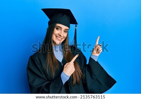 Beautiful brunette young woman wearing graduation cap and ceremony robe smiling and looking at the camera pointing with two hands and fingers to the side.  Royalty-Free Stock Photo #2135763115