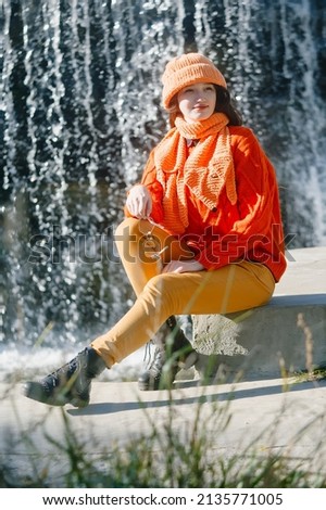 autumn portrait of a girl in a hat and orange sweater. woman sitting on the river bank