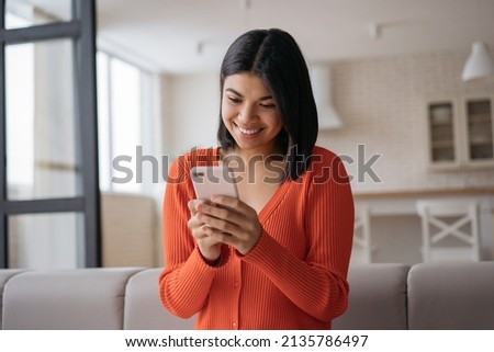 Young beautiful African American woman wearing casual clothing holding smartphone text messaging, communication online sitting at home. Smiling hipster female using mobile app shopping online