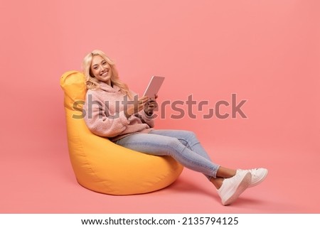 Happy lady using digital tablet while sitting in beanbag chair over pink background with free space. Relaxed woman reading book online or surfing internet