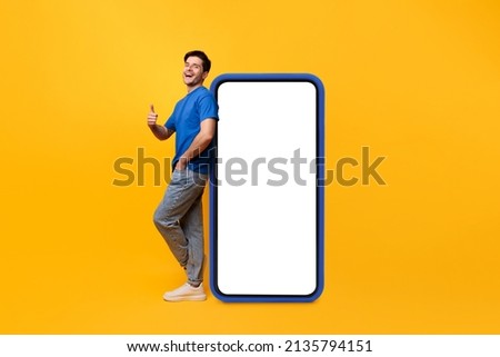Cool Mobile Offer. Excited casual guy leaning on big smartphone with blank empty white display, showing thumb up gesture, recommending great new app or website, mock up, full body length, yellow wall Royalty-Free Stock Photo #2135794151