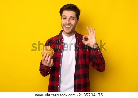 I Like Junk Food. Excited Guy Holding Showing Burger Gesturing Ok Sign Smiling To Camera Posing On Yellow Studio Background Wall. Young Man Approving Cheeseburger's Taste Eating Fastfood