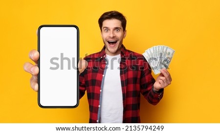 Cashback, Easy Online Money Transfer Concept. Excited young guy holding big smartphone with white blank empty cell screen for mock up and cash money dollar banknotes in hand, isolated on studio wall