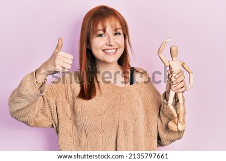 Redhead young woman holding small wooden manikin smiling happy and positive, thumb up doing excellent and approval sign 