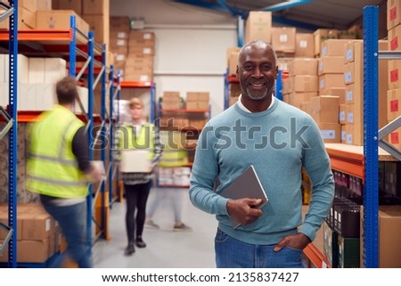 Portrait Of Manager With Digital Tablet In Busy Modern Warehouse With Staff In Background